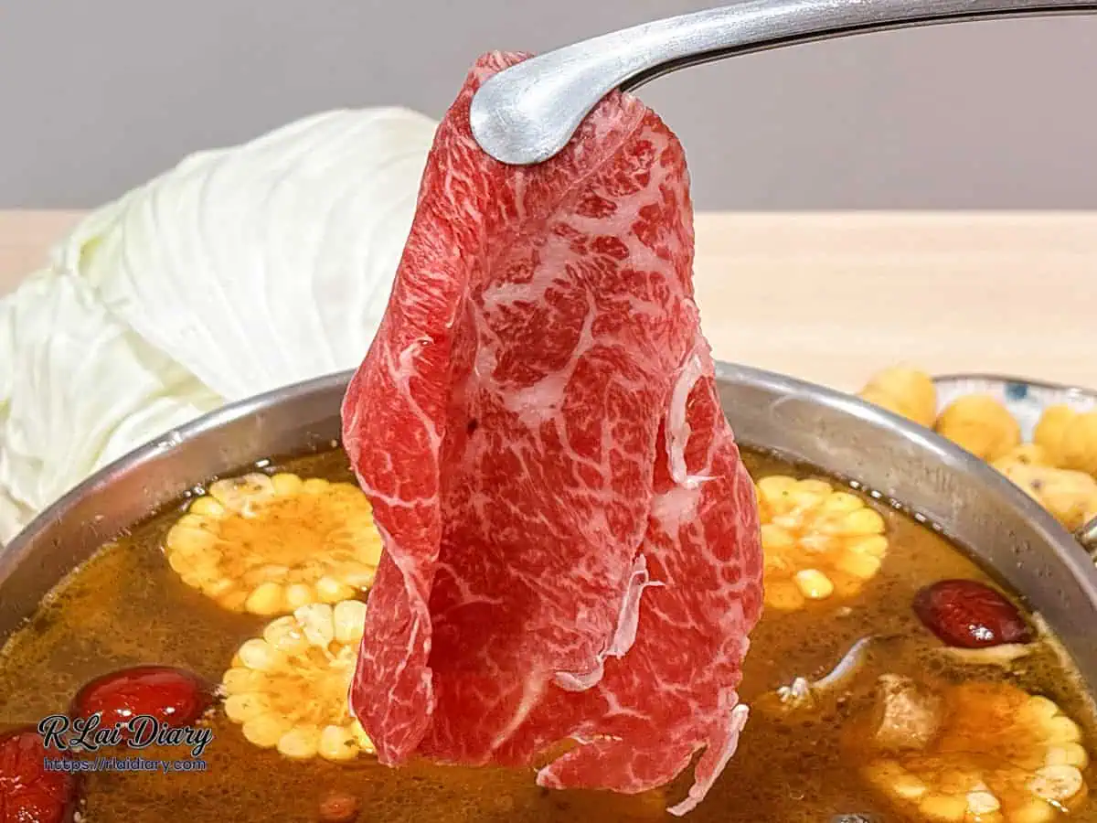 用夾子夾起大片的澳洲 M9 和牛放入麻油雞湯中涮煮，展示其誘人的油花分布