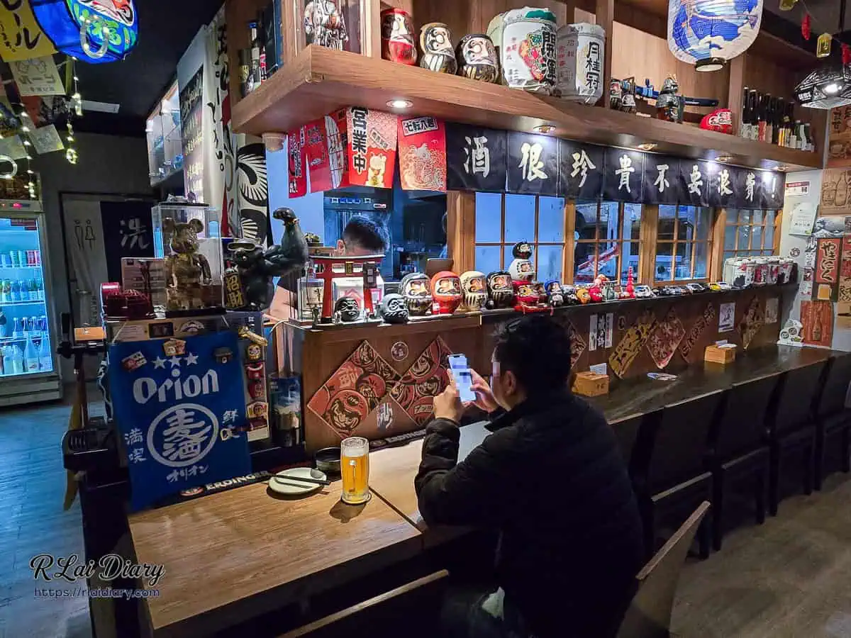 宇貳居酒屋吧台座位區，酒很珍貴不要浪費布條與大量達摩不倒翁裝飾